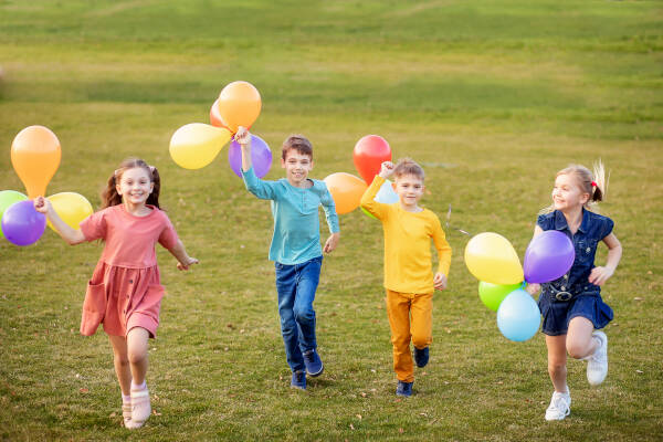 Four children running on a grassy field, each holding colorful balloons and smiling happily.