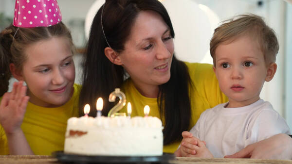 Mother sits at table with her daughter and two-year-old son, who prepares to blow out candles on his second birthday cake