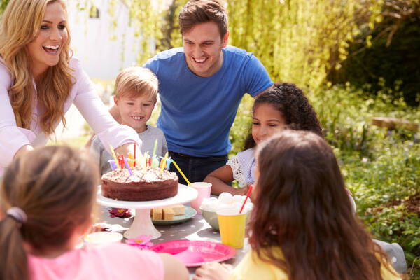 Parents and children enjoying a birthday party outdoors, with a mother presenting a chocolate cake with lit candles.