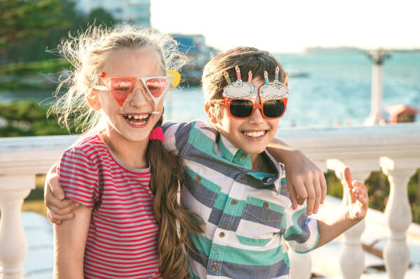 Two children wearing fun, oversized sunglasses and hugging each other at a birthday party by the seaside. Both are smiling and dressed casually.