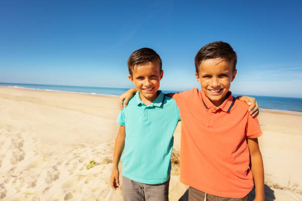 Two twin boys standing on a beach with their arms around each other, both smiling at the camera with the ocean in the background.