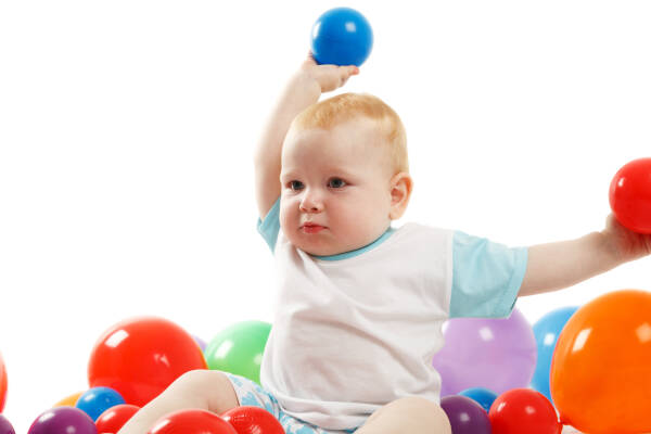 Two-year-old boy in a pool of colored balls, holding two balls in the air.