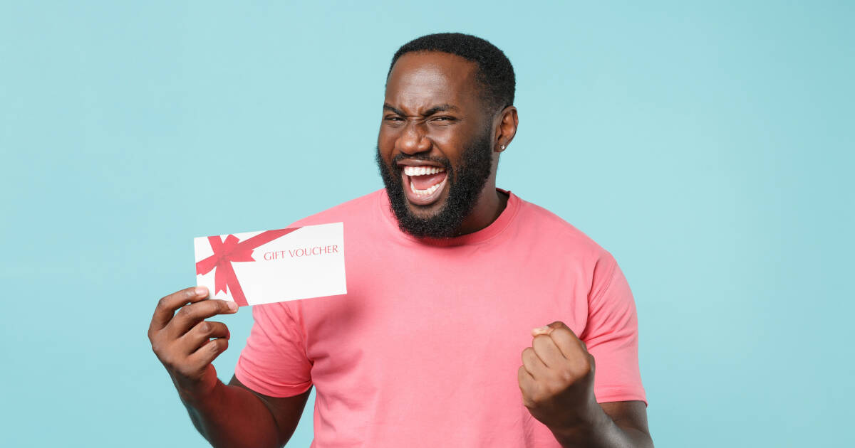 A joyful man in a pink t-shirt holds a gift voucher while celebrating with a big smile and a clenched fist, standing in front of a light blue background, the voucher features a red ribbon design.