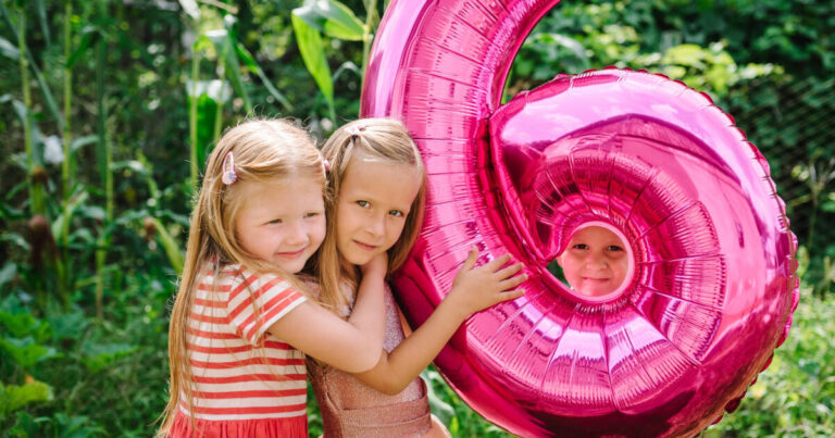 Three young children playing with a large pink balloon shaped like the number 6, boy peaks through the balloon.