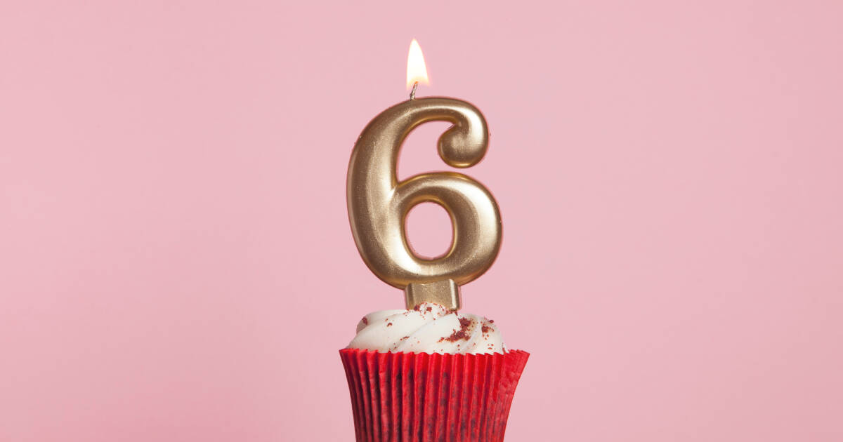 A cupcake with white frosting in a red wrapper, topped with a lit gold number 6 candle, set against a pink background.