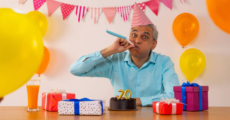 Man wearing party hat blowing party horn, sitting at a table with a 70th birthday cake and presents, surrounded by balloons.