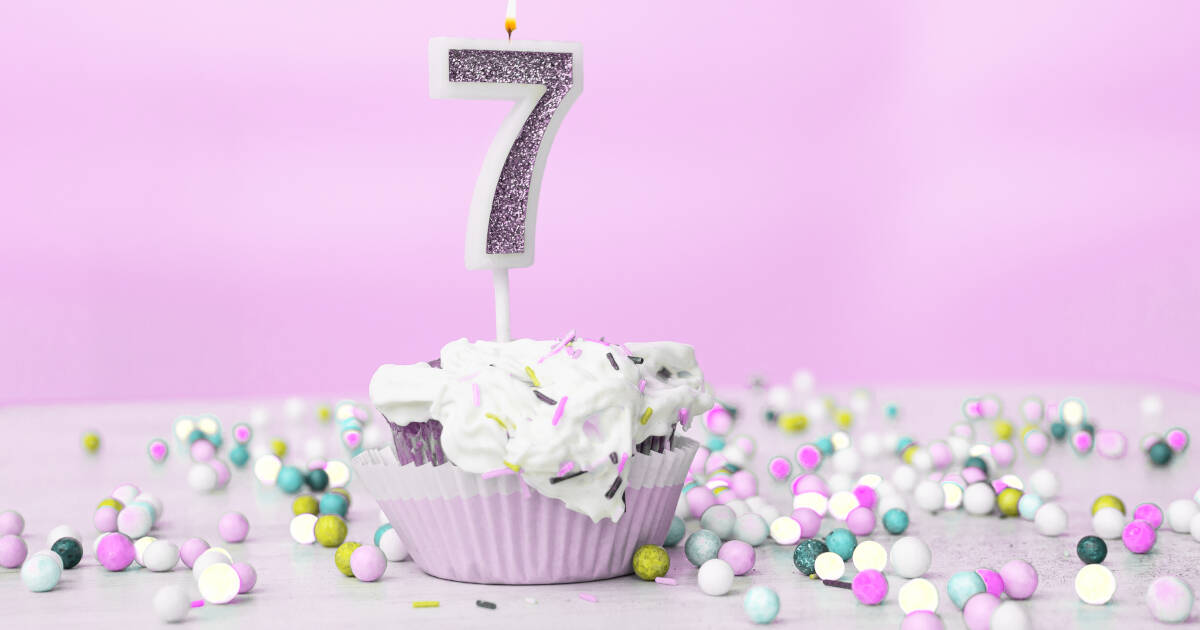 A cupcake, topped with a glittery number 7 candle, surrounded by colorful candy balls on a pink background.