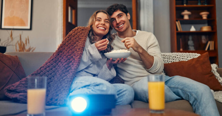 A smiling couple cuddled up on a cozy couch with a blanket, enjoying popcorn while watching a movie projected in their warmly lit living room.