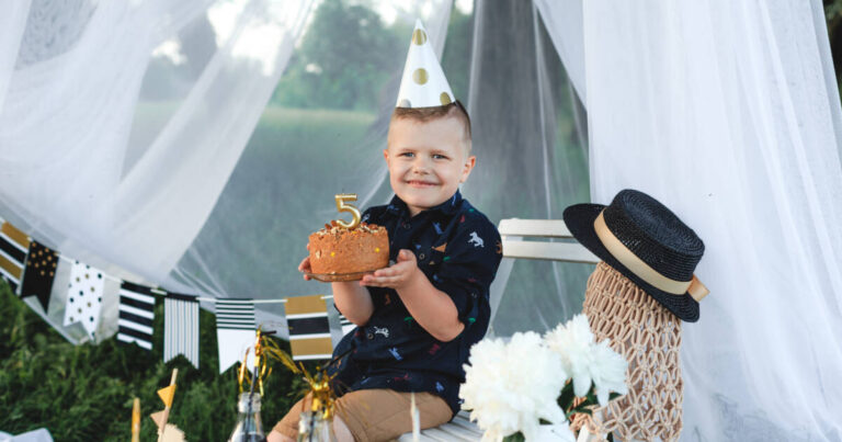 Smiling young boy wearing a party hat, holding a small birthday cake with a number 5 candle, celebrating outdoors.