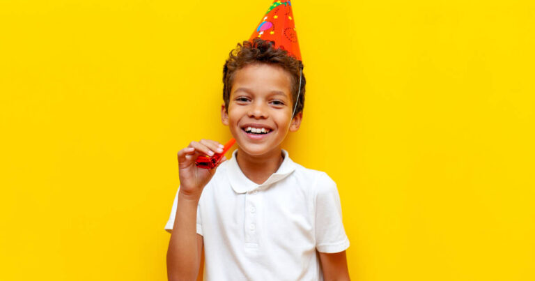 Young Boy In Festive red party Hat holding a party horn in his right hand in front of a yellow background,