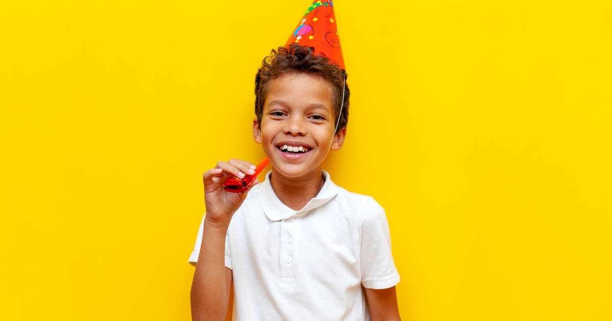 Young Boy In Festive red party Hat holding a party horn in his right hand in front of a yellow background,