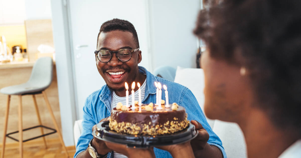 A man smiling brightly as he is presented with a chocolate birthday cake topped with lit candles in a cozy home setting.