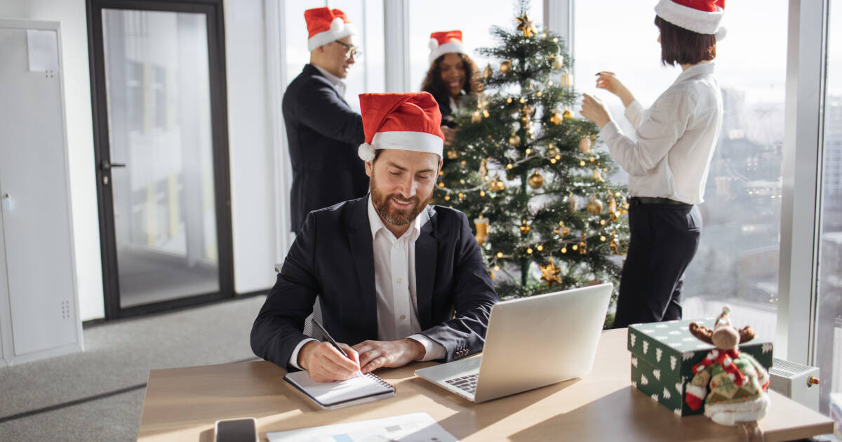 Businesspeople in an office wearing Santa hats, with one man working at his desk while others decorate a Christmas tree.