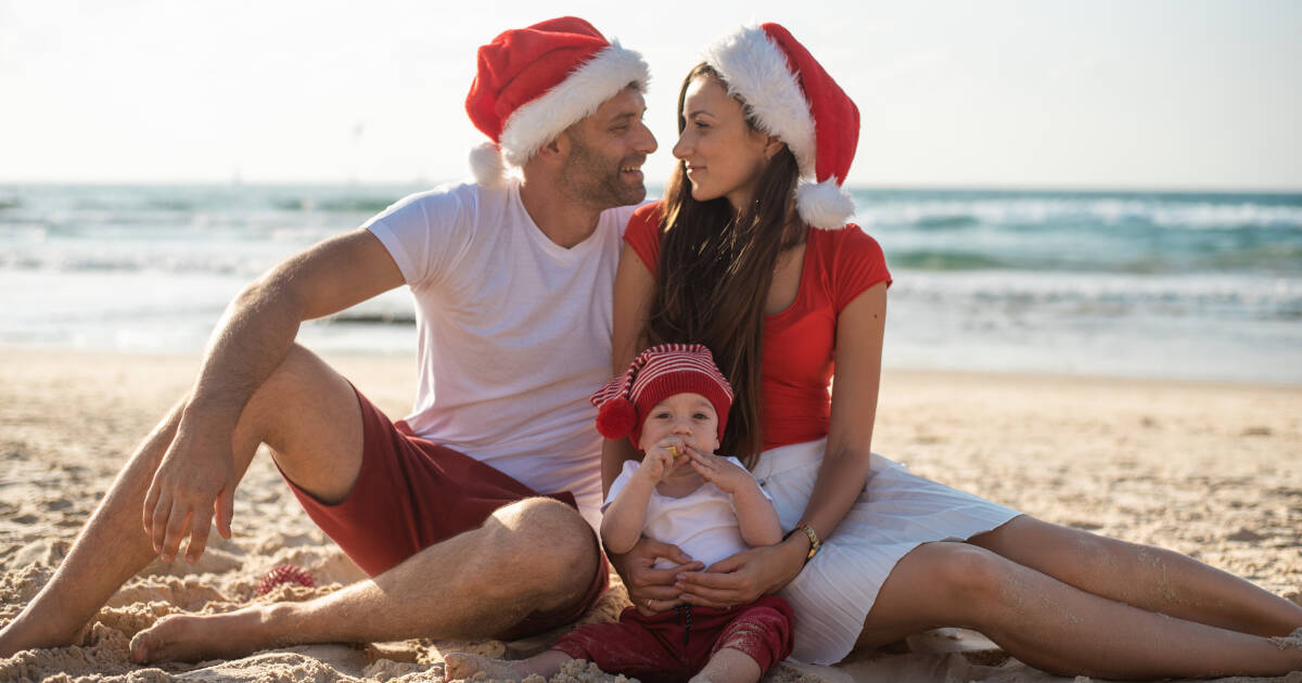 A family sitting on the beach, wearing Santa hats, with a father and mother smiling at each other while holding their baby.