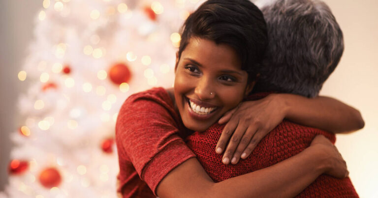 A woman smiling warmly while hugging her mother in front of a beautifully lit Christmas tree with red ornaments.