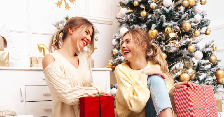 Two young women laughing and exchanging gifts while sitting next to a beautifully decorated Christmas tree.