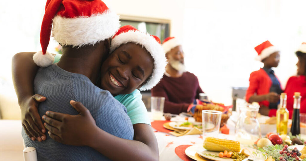 A joyful young boy wearing a Santa hat warmly hugs an adult, also in a Santa hat, they embrace with smiles, while a holiday meal and family members in Santa hats are seen in the background.