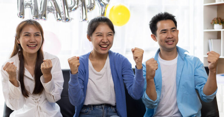 "Three cheerful young adults sitting on a couch celebrating, smiling brightly, and raising their fists in excitement, with a festive background including balloons and a 'HAPPY' banner."