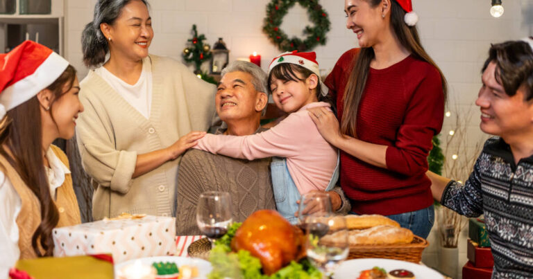 Family gathered around a holiday dinner table, with a young girl hugging her grandfather and everyone smiling.