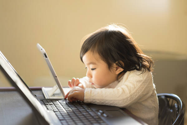 1-year-old girl plays on tablet beside larger laptop, appearing to 'work' in adorable imitation.