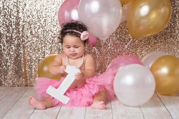 1-year-old girl plays with large number one block, wearing pink tutu, flower headband, pearls, with balloons.