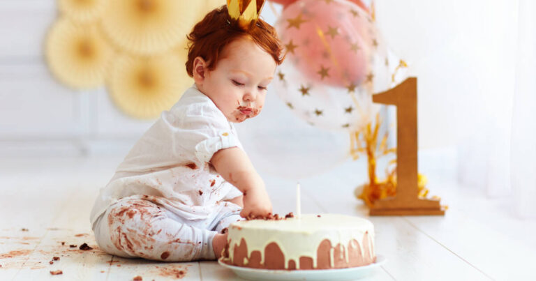 A curious baby boy pokes his finger into his first birthday cake with balloons and a number 1 in the background.