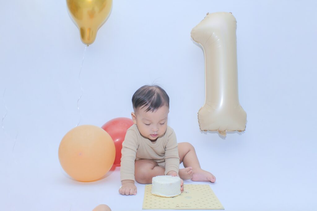 A baby boy sits next to a cake and balloons, with a number 1 balloon in the background, against a blueish-white backdrop.