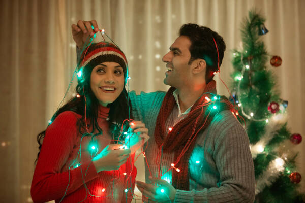 A cheerful couple tangled in festive Christmas lights, smiling as they decorate together.