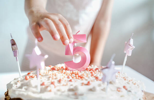 A child places a number 5 candle on a birthday cake decorated with white frosting, pink sprinkles, and star-shaped candles.