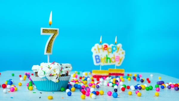 A colorful birthday cupcake with a golden number 7 candle, surrounded by rainbow sprinkles and a "Happy Birthday" sign.