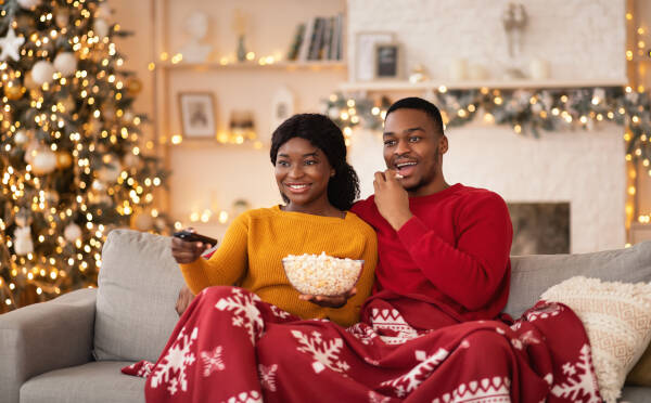 A couple cozy on a couch under red Christmas blankets, watching TV with popcorn in a festive living room.