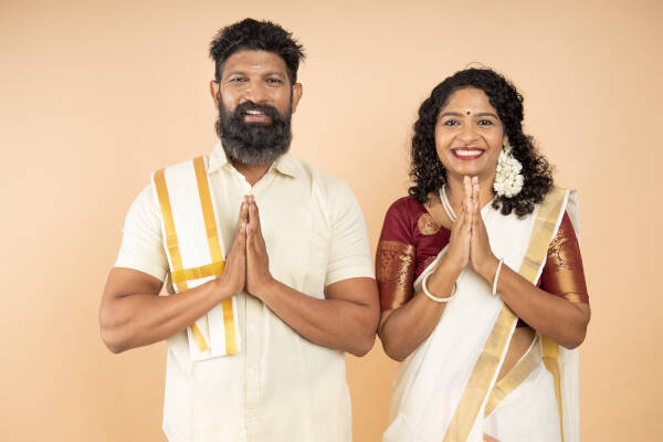 A couple dressed in traditional attire with hands pressed together in a greeting, smiling against a beige background.