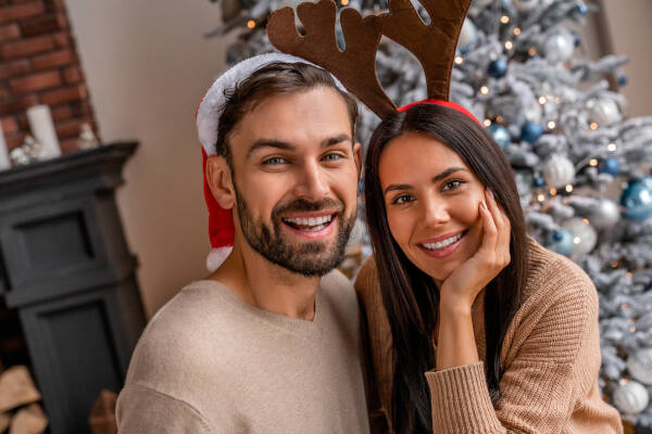 A couple wearing Christmas-themed accessories—Santa hat and reindeer antlers—smiling in front of a decorated tree.