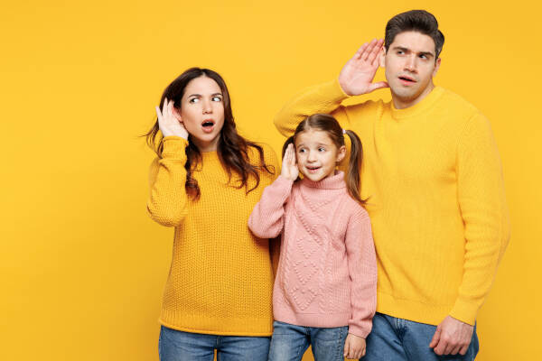 A family of three, all wearing matching colorful sweaters, posing playfully and cupping their ears against a bright yellow background.