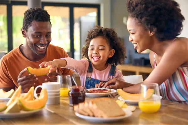 A family of three, including a young girl, sit at a table, smiling and sharing a meal together with fresh fruit.