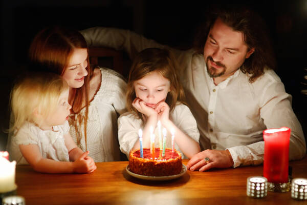 A family sits around a table lit by candles, watching a young girl as she prepares to blow out candles on her birthday cake.