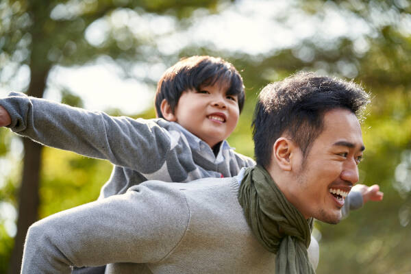 A father gives his son a piggyback ride in a sunny park, both smiling and pretending to fly with arms outstretched.