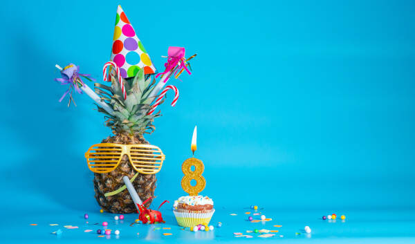 A festive pineapple wearing sunglasses and a birthday hat, next to a cupcake with a number eight candle on a blue background.