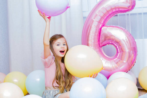 A girl surrounded by pastel balloons, holding one up with excitement. A large pink number 6 balloon stands behind her.