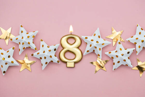 A golden number eight candle lit, surrounded by decorative white and gold stars on a pink background.