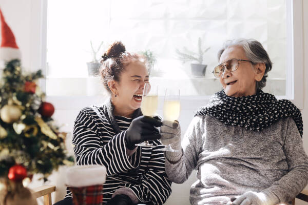 A grandmother and granddaughter clink glasses, laughing together near a Christmas tree, enjoying the moment.