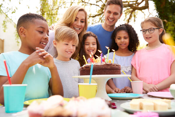 A group of kids at an outdoor birthday party, excitedly looking at a birthday cake with lit candles, surrounded by family.