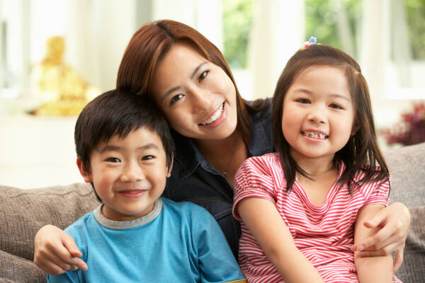 A happy mother sitting on a couch with her two smiling children, a young boy in blue and a young girl in pink stripes.