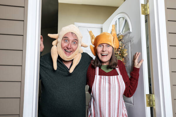 A man and woman in funny turkey hats, opening the door and smiling, celebrating a fun Thanksgiving moment.