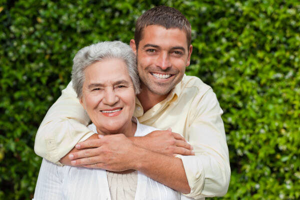 A man hugs his mother in the garden as they both smile at the camera.