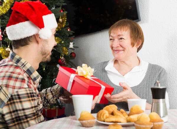 A man in a Santa hat gives a red gift box to an older woman near a Christmas tree, both smiling warmly.