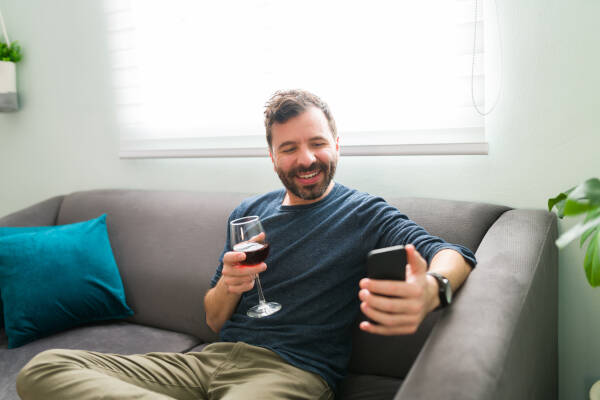 A man sitting on a couch, holding a glass of red wine, smiles while looking at his phone.