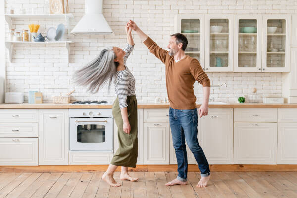 A middle-aged couple dances together in their kitchen. they share a lighthearted moment barefoot on the wooden floor.