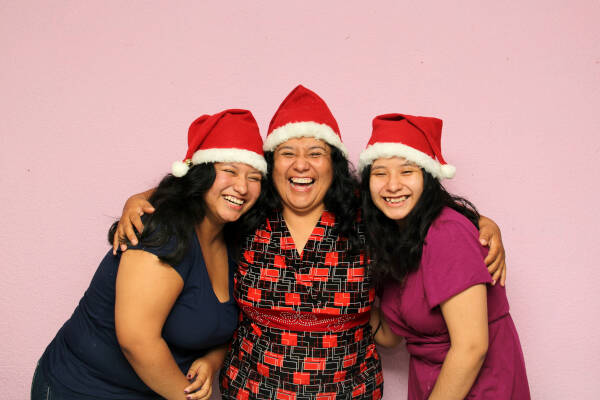 A mother and her two daughters in Santa hats laugh together, hugging in front of a plain pink wall.