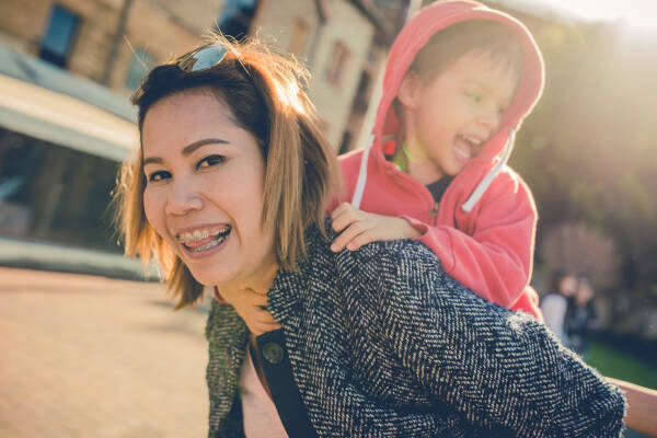 A mother carries her young child on her back as they both smile and laugh while enjoying time outdoors.